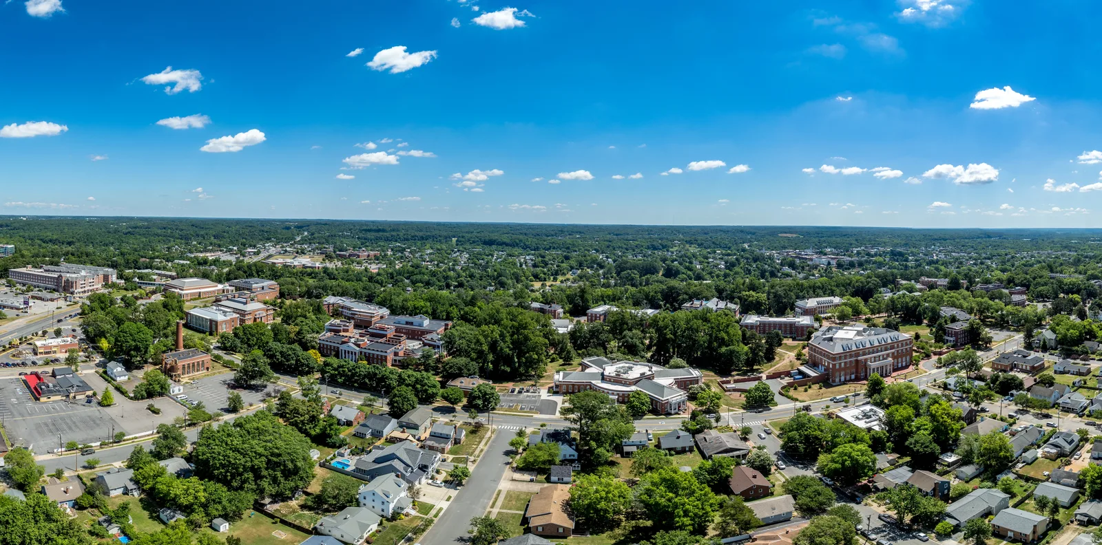 Aerial view of a suburban area with academic buildings, houses, roads, and trees under a blue sky.
