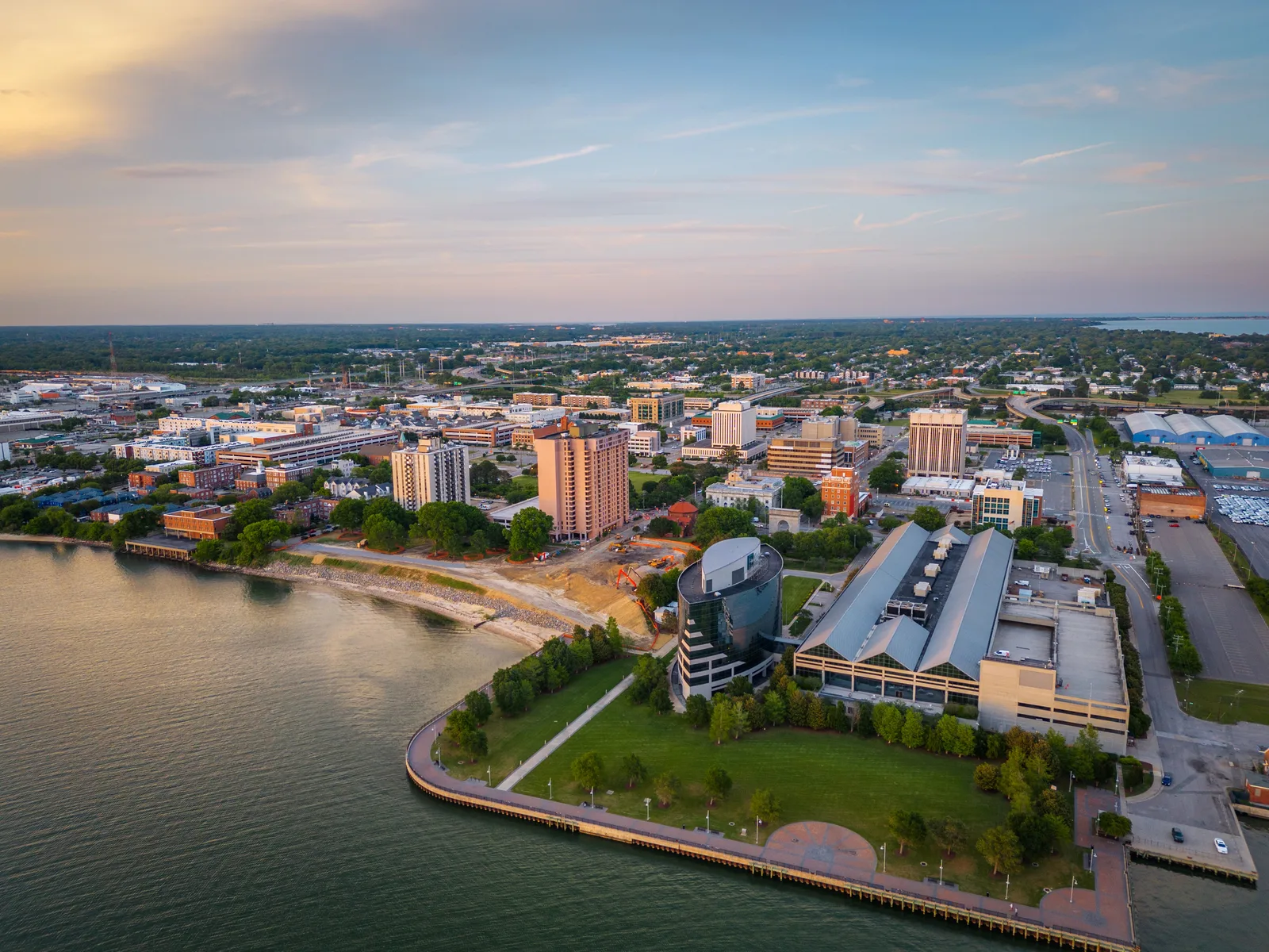 An aerial sunset view of the downtown Newport News waterfront, highlighting modern commercial buildings and green public parks stretching along the James River coastline.