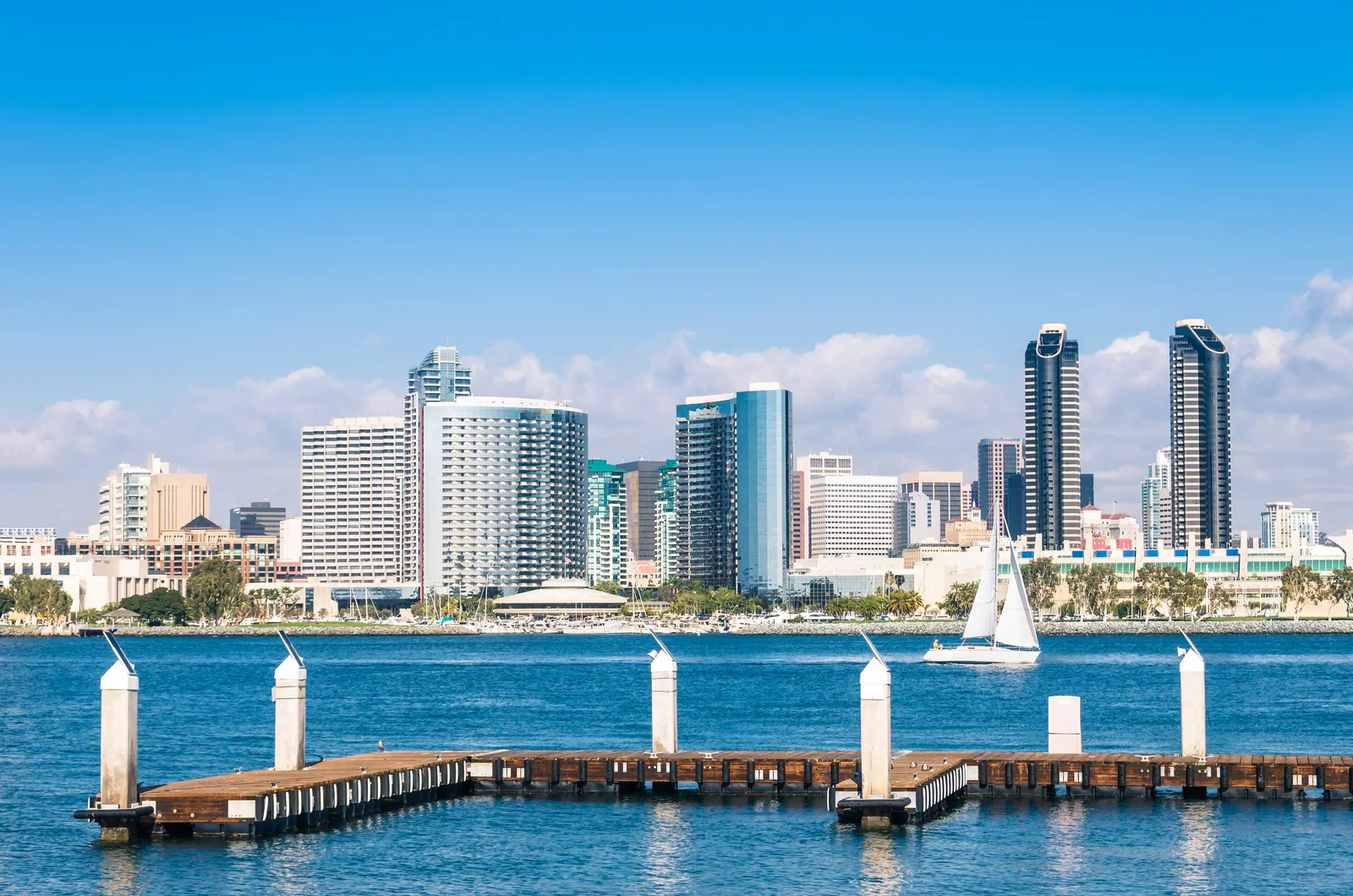 Downtown high-rise skyline across a blue bay, with a sailboat and empty wooden marina docks in the foreground