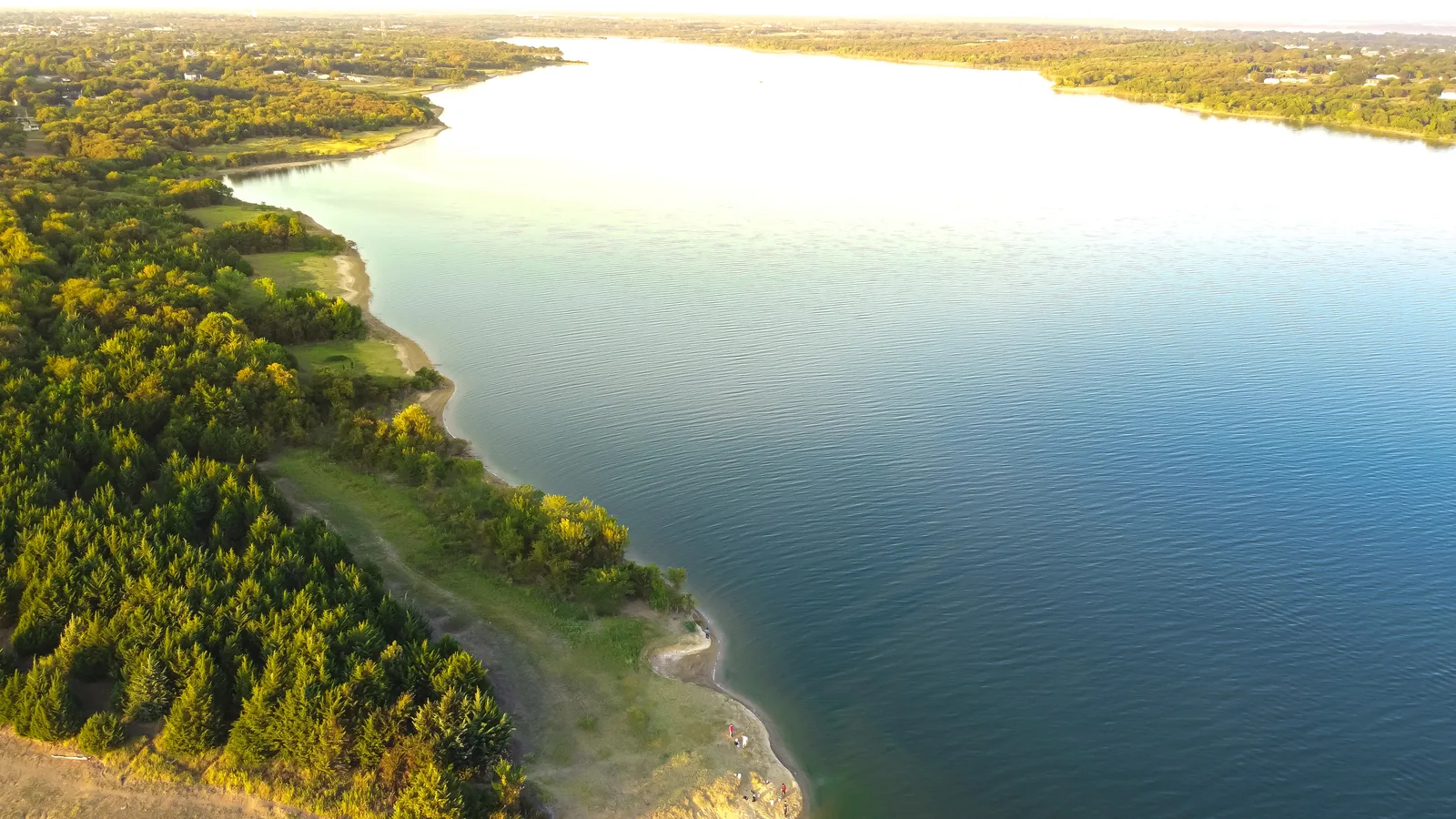 Aerial view of a broad lake with a wooded, sandy shoreline in low evening sun