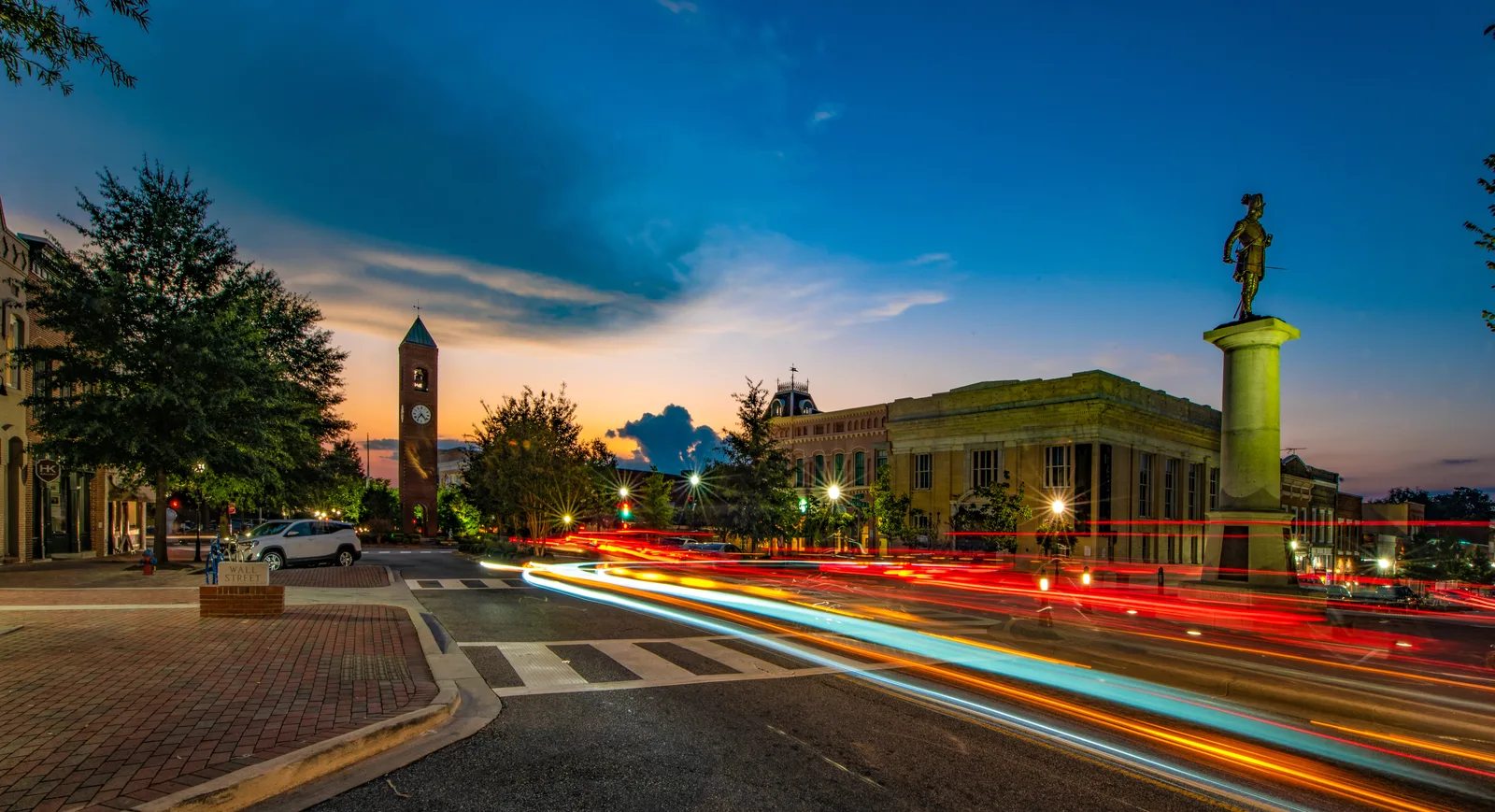 Downtown street at dusk with a brick clock tower, a statue on a column and light trails from passing traffic