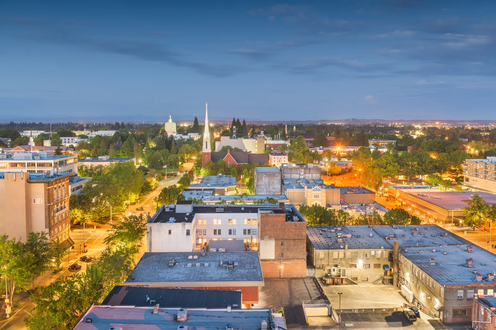 Rooftop view of a small city downtown at dusk, with a church spire and a domed building beyond tree-lined streets