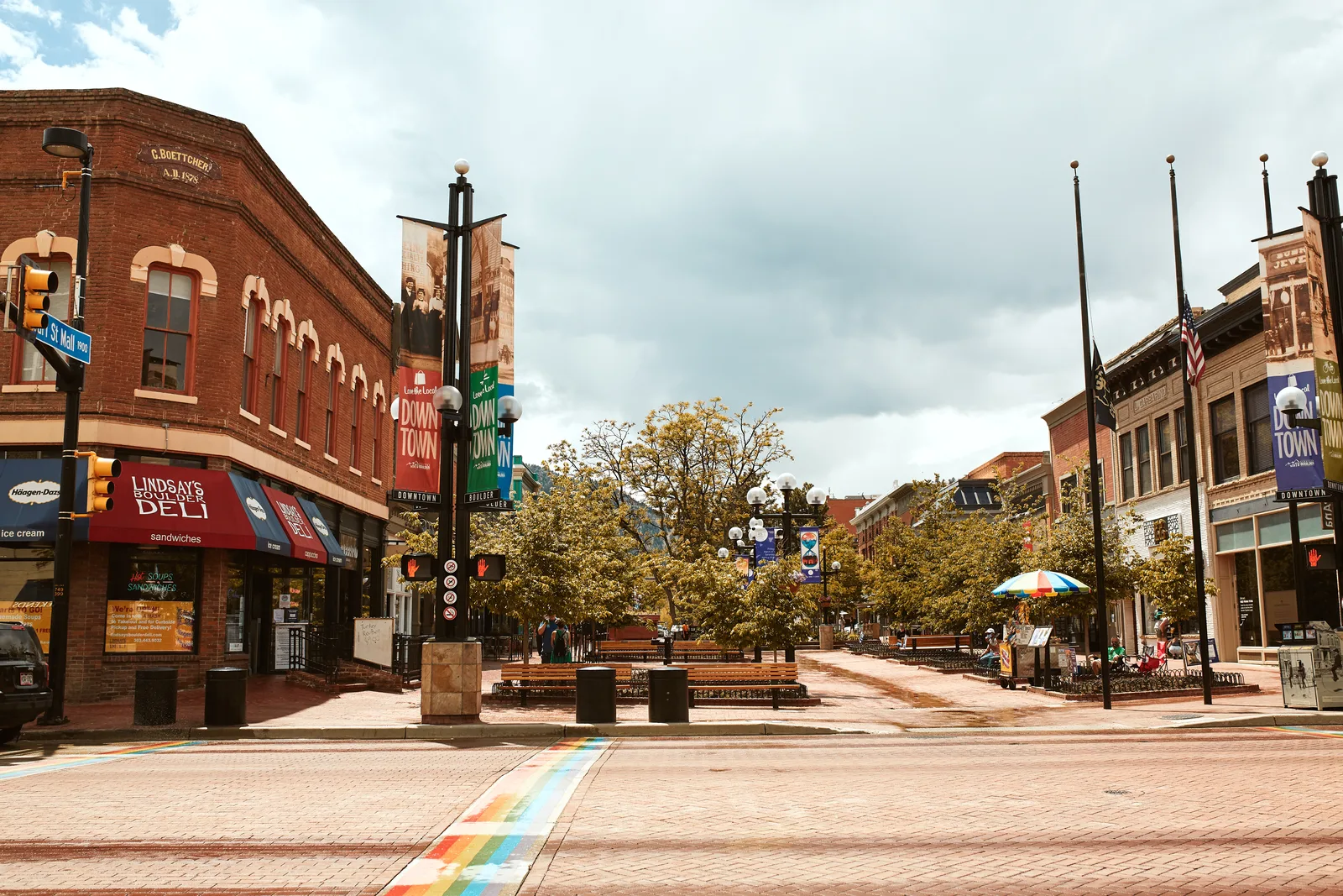 Brick pedestrian mall with a deli on the corner, benches, trees and banners reading Downtown Boulder