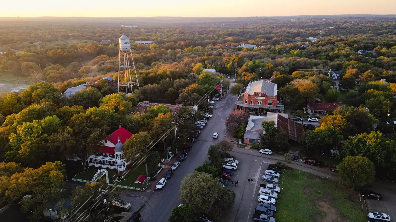 Aerial view of a small town street with a white water tower, historic brick buildings and parked cars among trees
