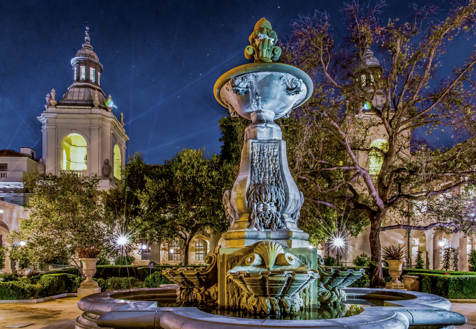 Ornate stone fountain lit at night in a formal courtyard, with a domed civic tower and trees behind