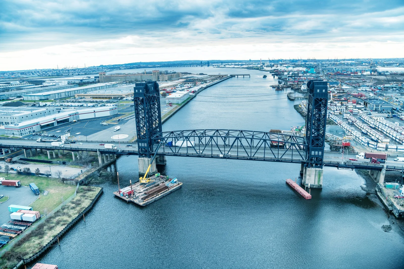 Aerial view of a steel vertical-lift bridge over a river, with a construction barge and industrial land on both banks