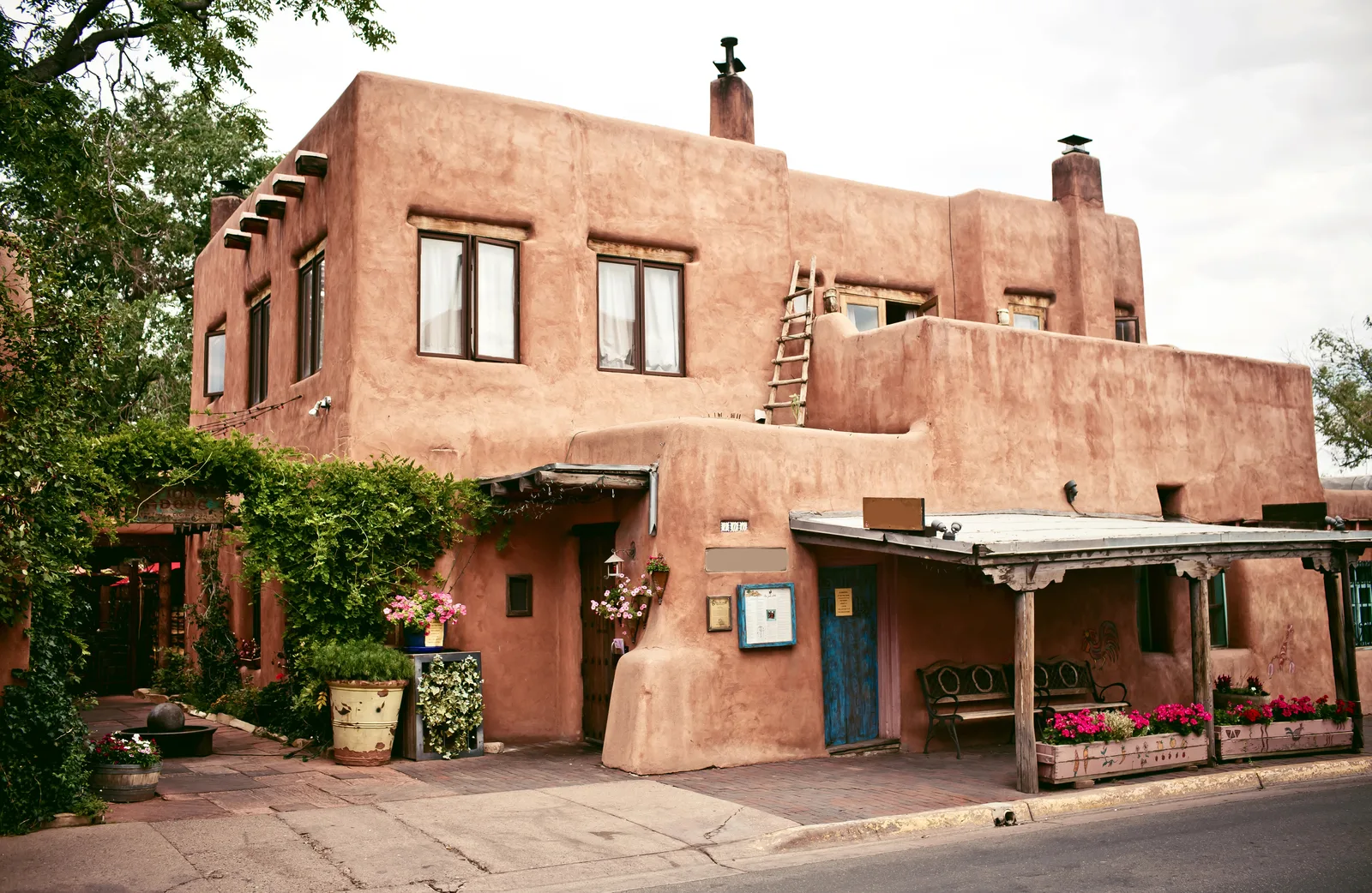 Two-storey adobe building with a wooden ladder, blue door, covered porch and flower boxes along a street