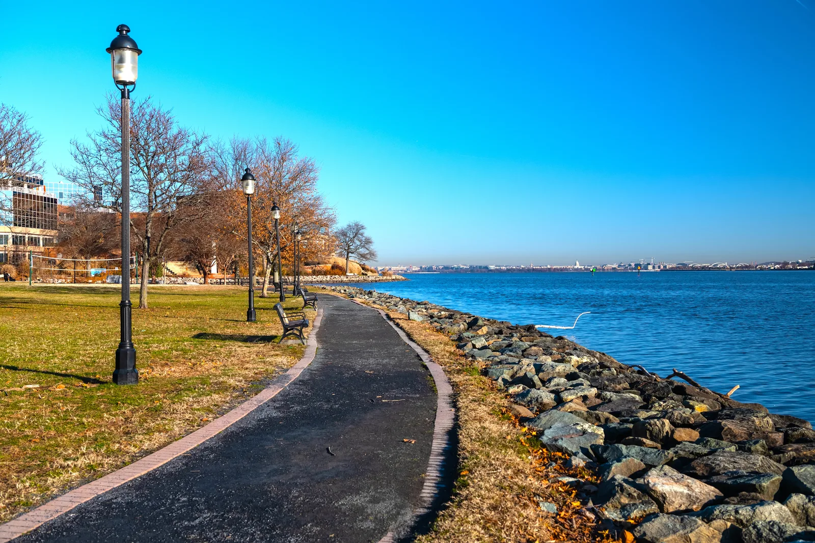 Paved riverside path with lampposts and benches beside a rock-lined shore, a city skyline across the water