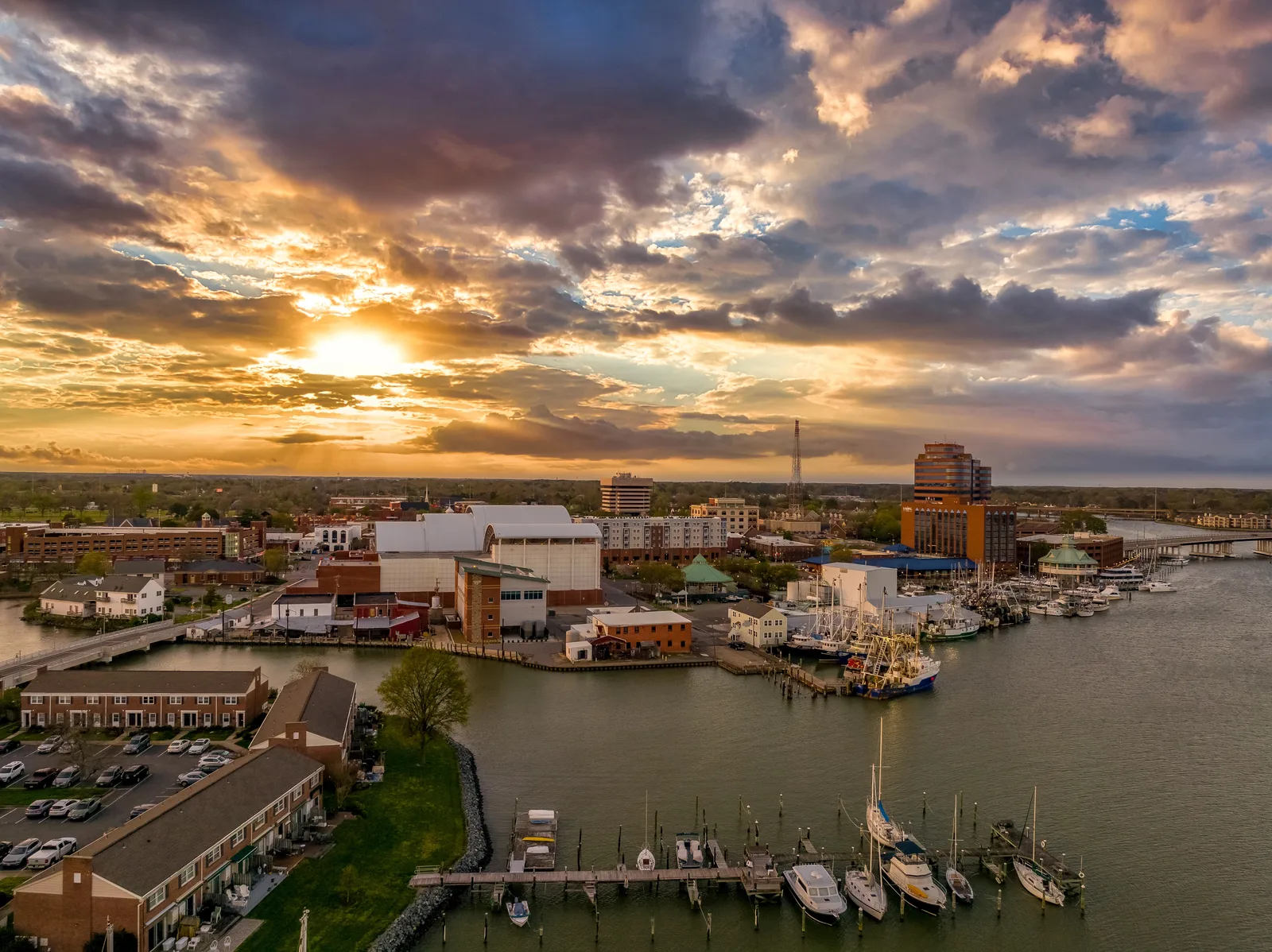 Aerial view of a waterfront town at sunset, with marinas, docked fishing boats, a bridge and mid-rise buildings