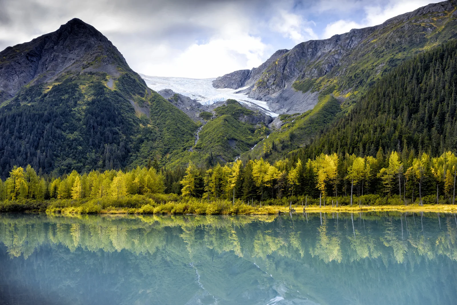 A blue glacier nestled between lush green mountains reflects clearly in a calm lake bordered by autumn-colored trees in Alaska.