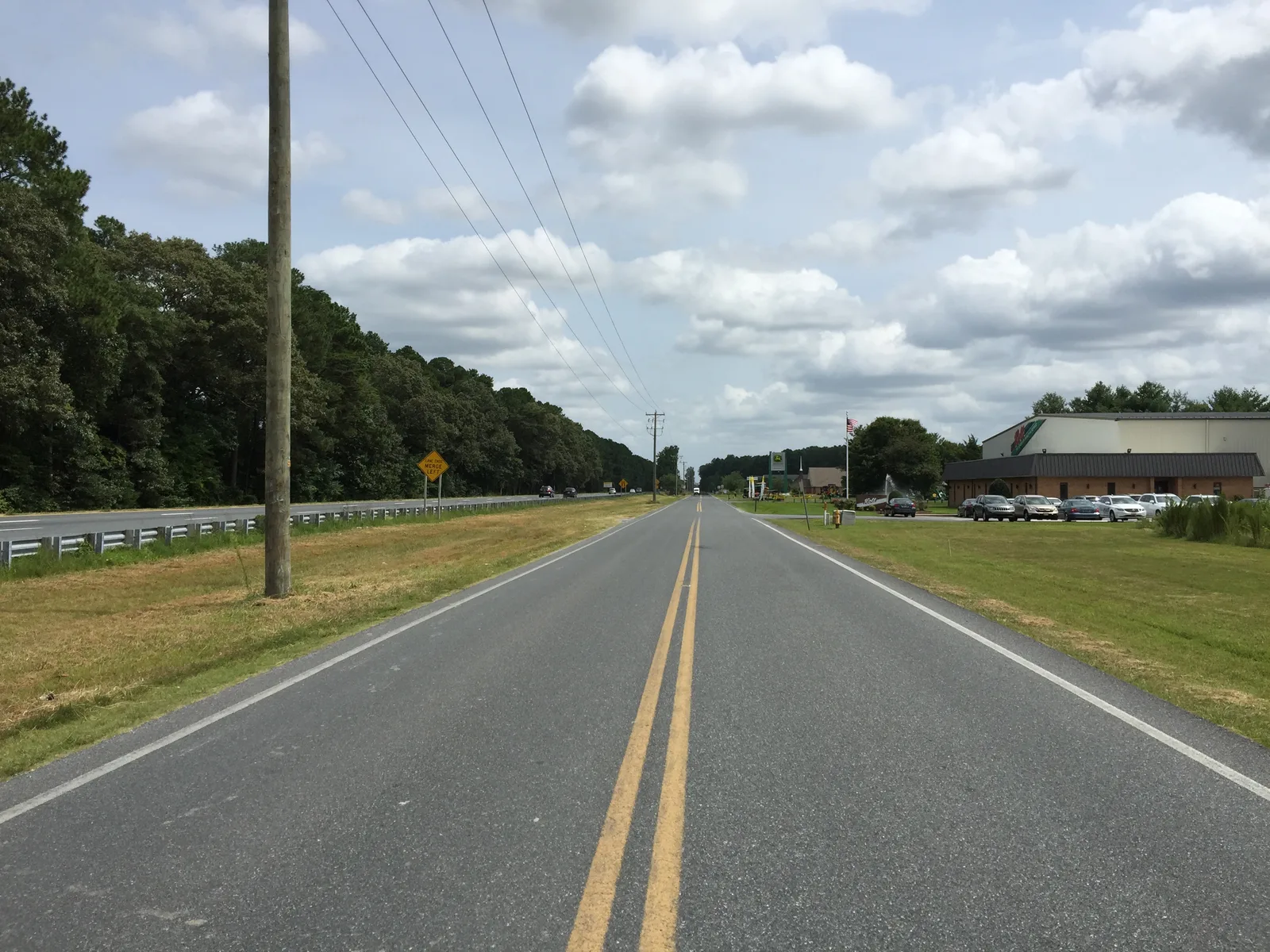 Two-lane rural road with double yellow lines beside a divided highway, utility poles and a commercial building