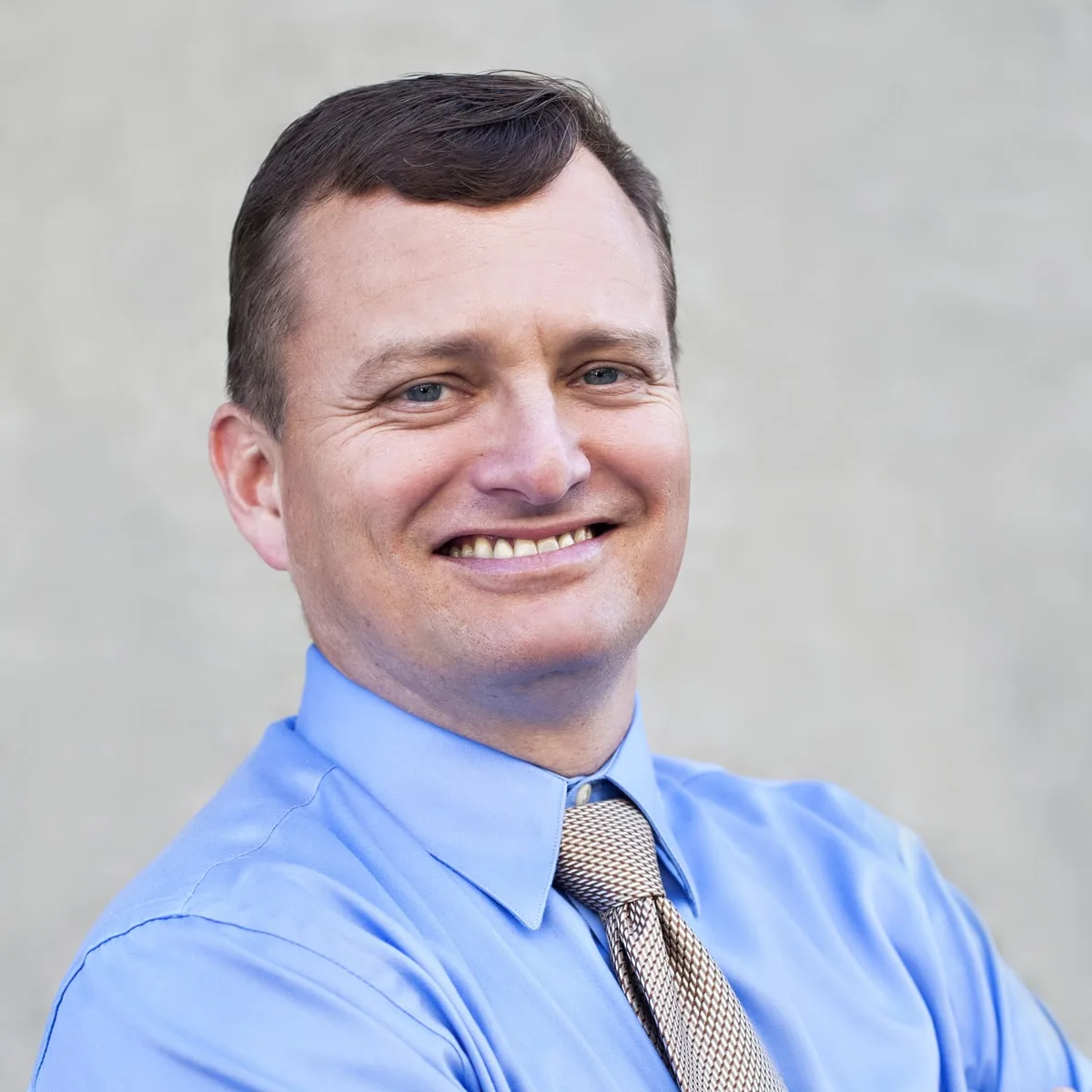 Headshot of a smiling man with short dark hair in a blue shirt and patterned tie, grey background
