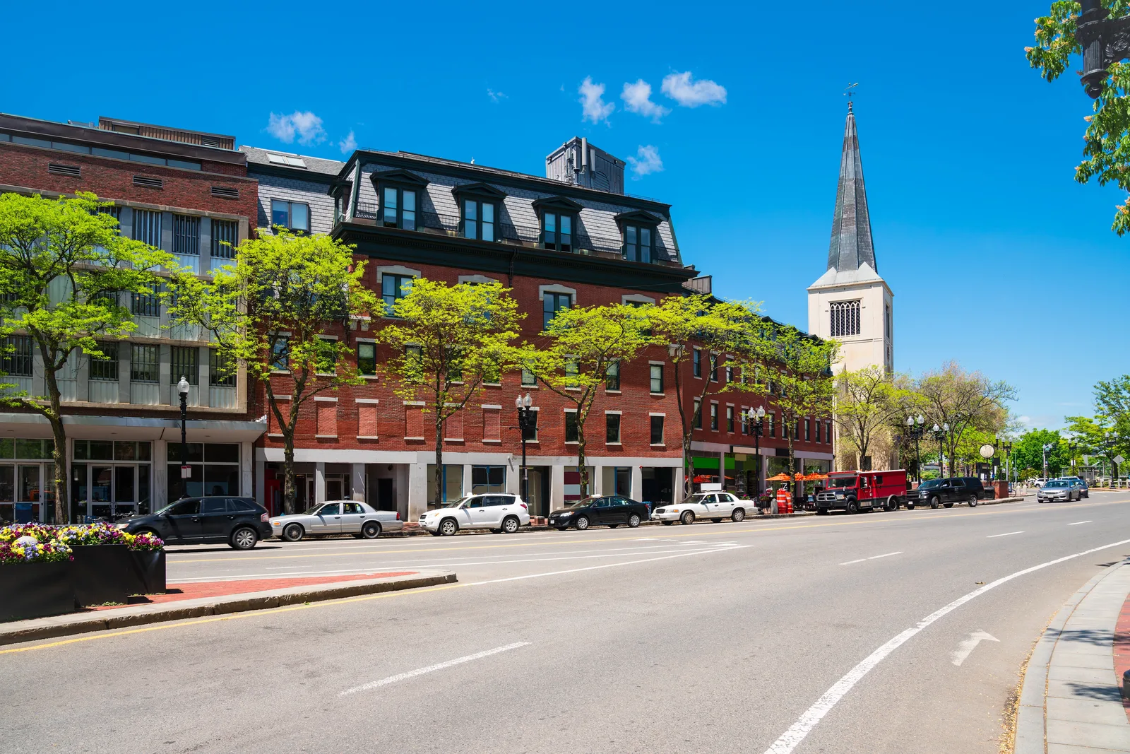 Tree-lined city street with red brick mixed-use buildings, parked cars and a white church steeple