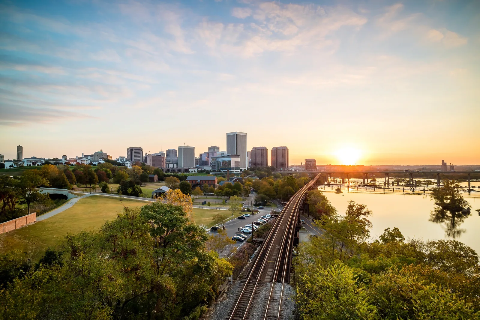 City skyline at sunrise beyond a park, with an elevated rail line running beside a wide, rocky river