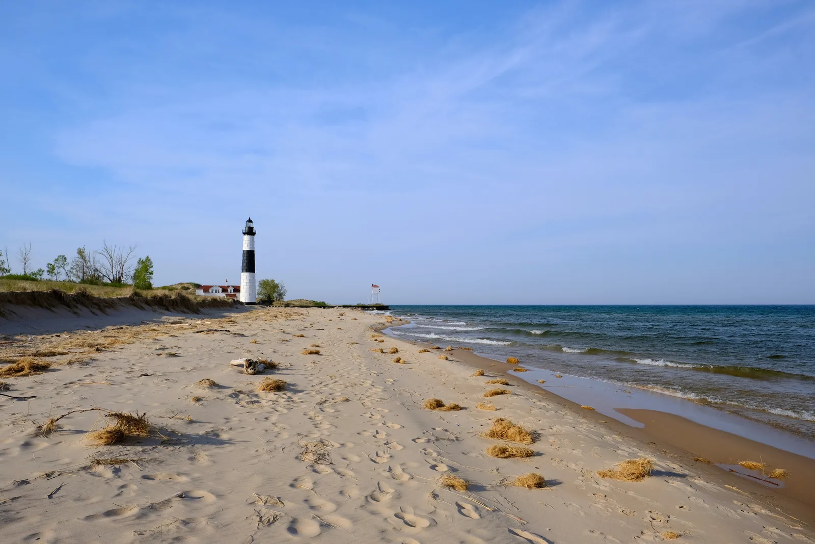 Black-and-white striped lighthouse behind sand dunes on a wide beach beside open blue water