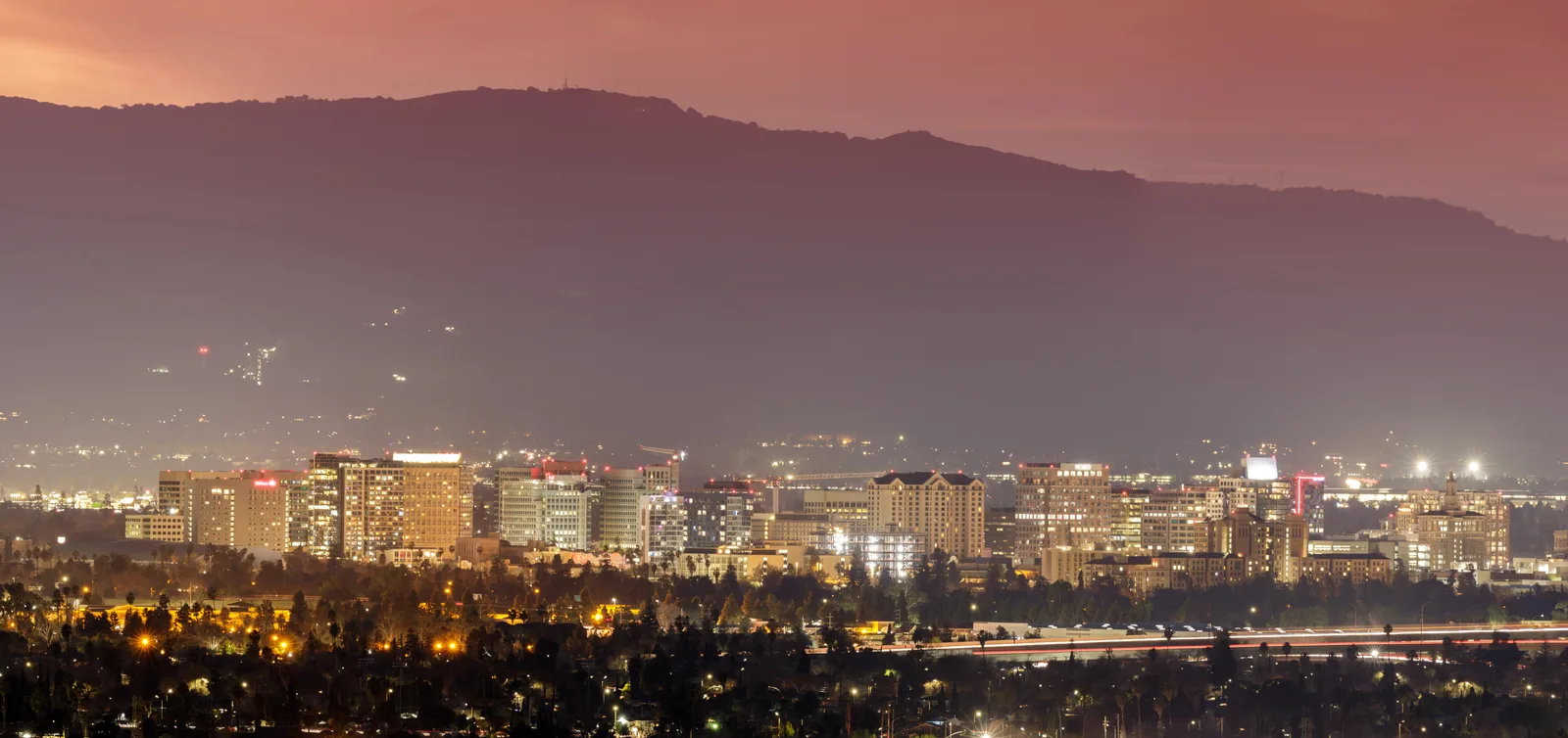 City skyline of mid-rise towers lit at dusk, with a dark mountain ridge behind under a pink sky