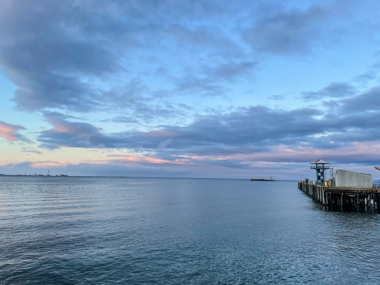 Calm harbour water at dusk under blue and pink clouds, with a wooden pier and a barge on the horizon
